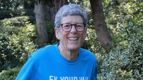 A woman with short grey hair and round glasses wearing a blue T-shirt runs in a park