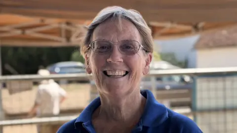 A picture of Elizabeth Miller, who is wearing a blue T-shirt and glasses. She has short, blonde hair.