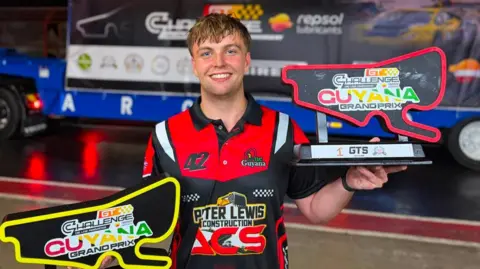 A man in a red and black T-Shirt smiling at the camera holding two trophies in the shape of a race track.