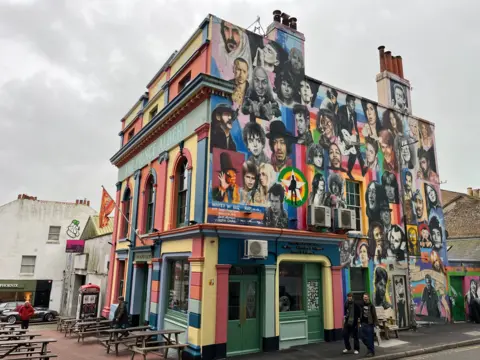 George Carden/BBC The Prince Albert building which is multicoloured in yellow, blue, green, orange and red paint. One wall is covered with a mural of celebrities including Elvis Presley, Eminem and Amy Winehouse. There is also a Banksy piece of art called Kissing Coppers. There is a large Prince Albert logo on the front and wooden benches outside the front and a flag hanging down