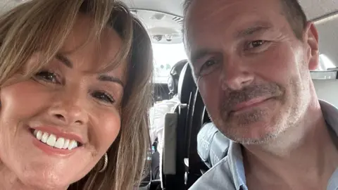 Ross Kaye A selfie of Ross and his wife Tracey. Tracey has short brown hair and a fringe. She is smiling at the camera. Ross has short brown hair and facial hair, he is wearing a blue shirt and is smiling at the camera. They are sat inside what looks like a plane.