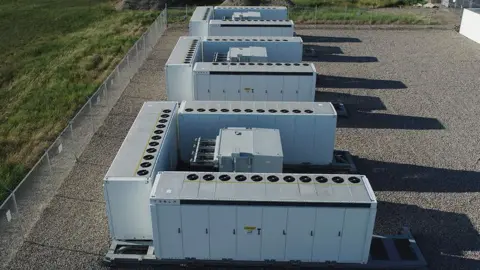 A general view of part of a battery storage facility showing three U-shaped white buildings with doors down the sides, on a gravel base, surrounded by a tall wire fence and grass