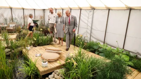 PA Media King Charles III and Queen Camilla are stood with two other people on wooden decking which is surrounded by different plants. The garden is in a white marquee tent. 