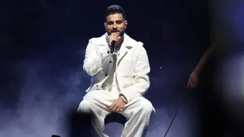 Getty Images A man wearing all white sitting on stage with a mic in his hand, wearing all white. The background is dark and smoky.