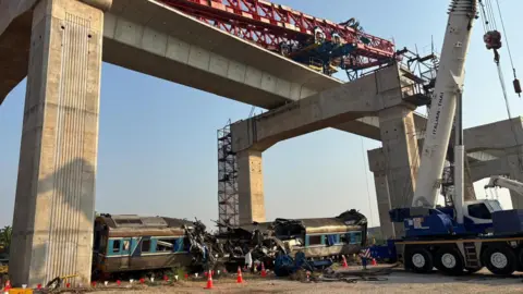 A crane lying sideways along the overhead rail track and a wrecked blue train on the ground below. Some of the train's crashes are completely crushed