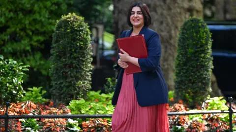 Bloomberg/Getty Images Shabana Mahmood, UK home secretary, arrives for a cabinet meeting at 10 Downing Street in London in a pink dress with a navy blazer, while holding a red box and smiling. Green foliage is seen behind her.
