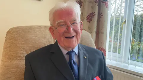 BBC William Wake is laughing. He is a man in his 90s with white hair and glasses and is wearing a suit and tie and a pale blue shirt.