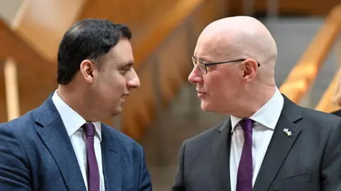 Anas Sarwar, who has short dark hair and is wearing a blue suit, white shirt and tie, is face to face with John Swinney who is bald and is wearing glasses, a dark grey jacket, white shirt and purple tie