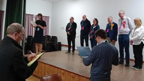 Political candidates gathered on a stage as results are read out. Each candidate is wearing a rosette. In the foreground a man is writing on a clipboard and another man is holding open a laptop.