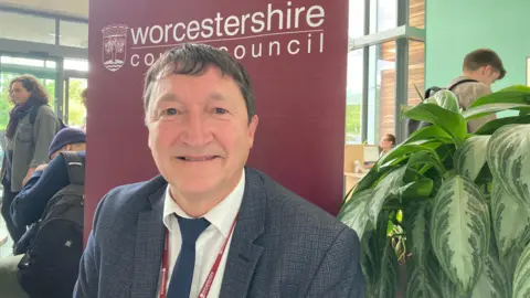 Mark Fitton, a Worcestershire County Council director, is sat down at a table with a large red banner behind him, which has the council's logo on it.