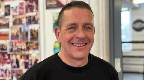 Simon Thake/BBC A man with shortly cropped dark hair and a black jumper smiles at the camera. Behind him are lots of photos pinned to the wall of people boxing.