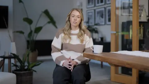 A blonde woman wearing a brown and white jumper with wide stripes and black trousers. She is sitting next to a table on top of which is a white mug