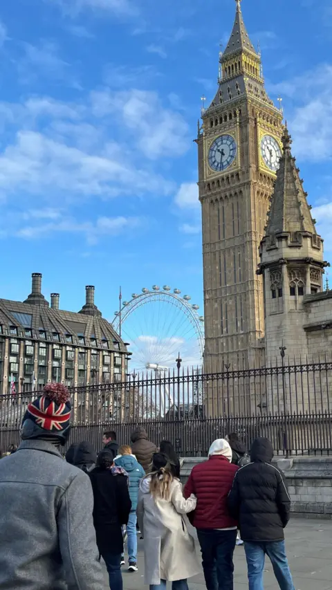 Tourists, including one wearing a union jack hat, crossing the road towards Big Ben, with the London Eye visible in the distance