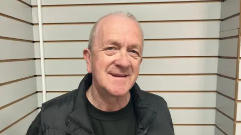A man with thinning white hair looks at the camera with a slight open-mouthed smile. He wears a black jacket over a dark top and is in front of walls with thick white stripes broken up by thinner brown ones.
