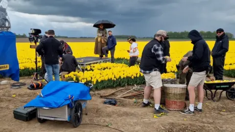 Mark Eves Film crew in a field of yellow tulips, surrounded by various filming equipment including a camera and small trailer. A woman in period costume is in the centre of the image, holding an umbrella