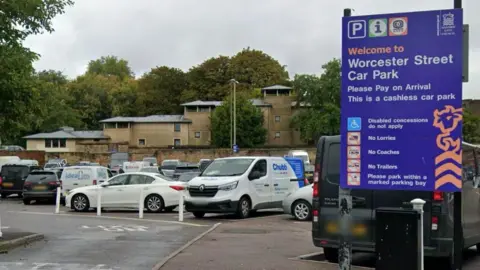 A large blue sign for the Worcester Street car park, with cars and vans parked behind it.