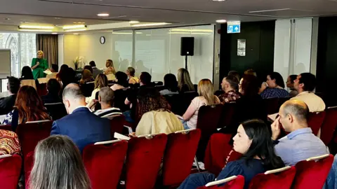 BBC/Nicola Rees A woman with blonde hair and a green dress is standing at the front of a hotel meeting room, talking to a crowd of people who are sitting on red chairs and looking forward.