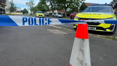 A residential street that has been cordoned off by police. In the foreground, there is a red and white traffic cone with blue and white tape stretched across it, clearly marked with the word “POLICE”. Behind the tape, at least two marked police vehicles are parked on the road, with one facing towards the camera and another further down the street. 