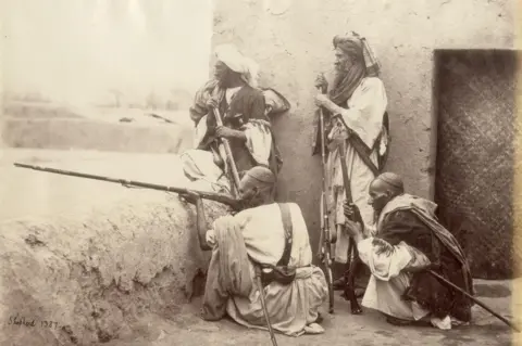 DAG Four Afghan tribesmen, armed with guns and dressed in traditional attire, photographed in 1862. The group - Afridis from the Khyber Pass near Peshawar - stand posed before the camera.
