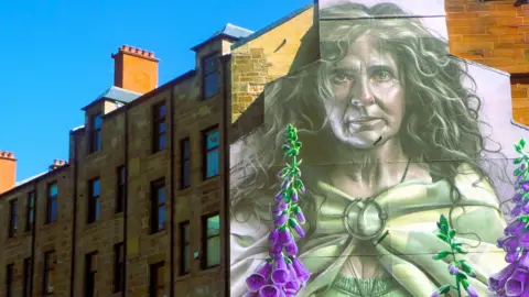 A mural of an older woman with long flowing grey hair, wearing a green shawl with a large brooch. She is surrounded by purple flowers. The mural is painted on the gable end of a sandstone tenement building.