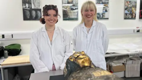 The students stand by Pa-Sheri who is resting on a table.  From this angle the gold of his mask can be seen.  The students wear white lab coats.
