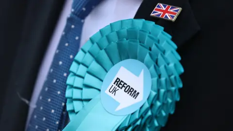Light blue Reform Party rosette positioned on a person wearing a dark suit with a blue shirt and darker blue tie. A union jack pin badge is positioned above the rosette.