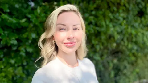 Mark Norman BBC A woman half smiles at the camera, she is wearing a white top with a green hedge behind