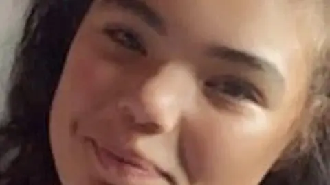 A close-up of the face of a young woman. She has dark curly hair, and is smiling at the camera