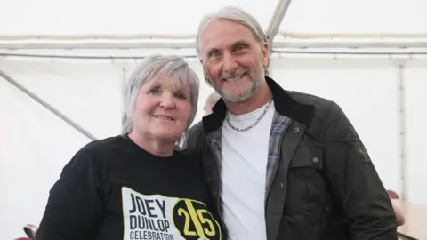 Pacemaker Linda, left, has short grey hair and is smiling at the camera. She is wearing a black t-shirt which says Joey Dunlop Celebration with the number 25 in yellow. She is standing next to Carl Fogarty. They are hugging into each other. Carl has longish grey hair pushed behind his ears. He is wearing a green coat with blue tartan lining and a white t-shirt. He is smiling at the camera.