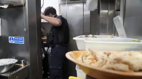 A kitchen where there are bowls of food in white plastic containers and a wooden bowl on the left side on a counter and then an immigration officer standing at the doorway of the kitchen leaning into another area where you can see the elbow and t-shirt of the person who is he speaking to. On the left, in the foreground is a metal sink with the sign 'now wash your hands' above it.