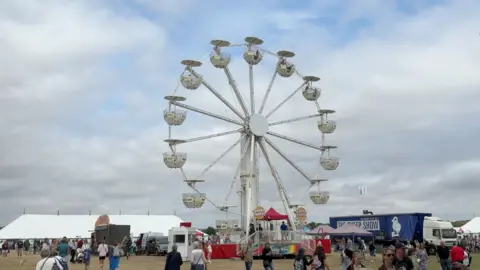 A 98ft-high (30m) big wheel. People are walking on the grass near it. The sky is blue and cloudy.
