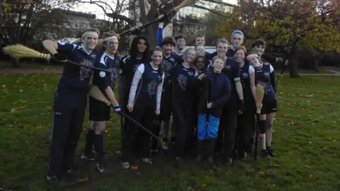 Sam A group of people stand in front of a tree. Some hold broomsticks, others wear headbands. A young boy in blue trousers stands in the centre of the group, smiling.