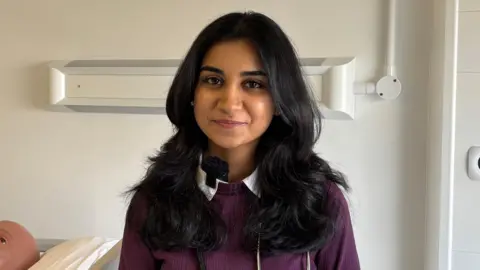 A woman with long brown curly hair smiling in the camera. She is wearing a maroon jumper with a while shirt with collars and has a stethoscope round her neck. 