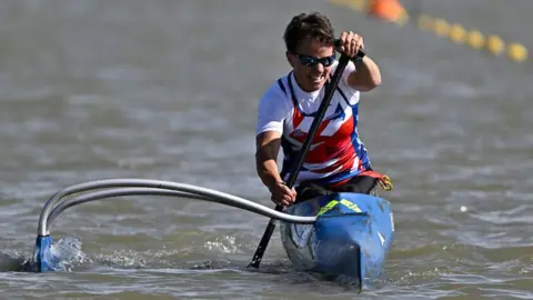 Inclusive Sport & Culture Festival An athlete with short brown and wearing sunglasses and a white, red and blue t-shirt is rowing in a canoe.