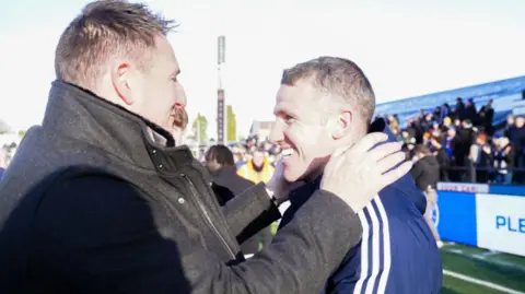 PA Chairman Rob Smethurst, wearing a dark jacket, puts his hand on his smiling manager John Rooney who is wearing a blue club jacket