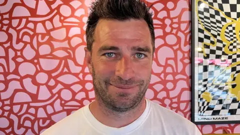 Peter Davis is a middle-aged man is standing in front of a wall covered in modern pink and red flowers. It also has a print on it that is black and white checks with a yellow scribble. He has short spiky dark hair and, dark eyebrows, blue eyes and dark stubble. He is looking straight at the camera and smiling. He is wearing a white T-shirt with a blue pattern in the middle.