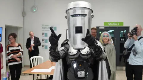 Count Binface arrives at the Clacton by-election vote count as people watch. He is pointing dramatically ahead of himself.