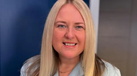 Head-and-shoulders portrait of Lesley with long light-coloured hair, wearing a light blue collared top and a thin necklace, looking toward the camera against a softly blurred indoor background with dark blue and neutral tones.