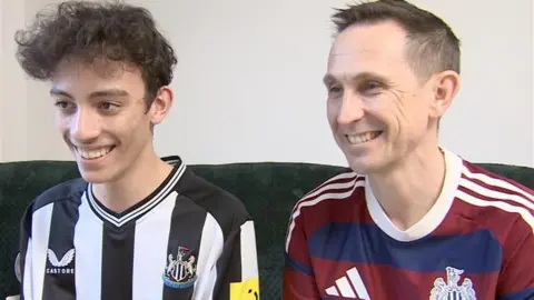 Charlie and Peter Maguire sitting adjacent  to each   different   successful  antithetic  Newcastle United tops connected  a sofa. Charlie has dark, curly hairsbreadth  and is wearing the achromatic  and achromatic  location  kit. Peter is wearing the reddish  and bluish  striped distant  kit. Both are smiling and looking distant  from the camera.