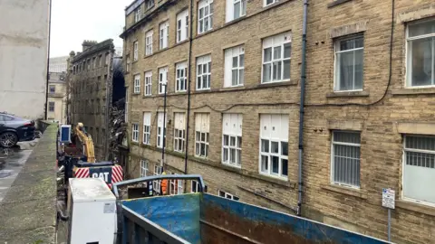 A battered blue skip in the foreground and a Yorkshire stone building with a collapsed section running from roof level to the first floor.