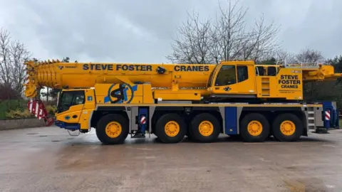 MANX UTILITIES A large mobile crane. It is a long, yellow vehicle with five sets of wheels and Steve Foster Cranes written in black lettering on the side.