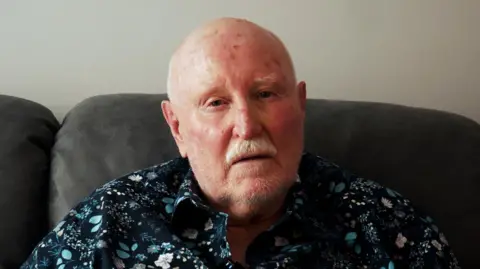 BBC Sam looking into the camera. He's sitting on a dark grey sofa wearing a blue floral shirt