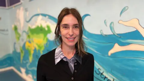 Viviana Durante posing against a mural on the sports hall wall. She's wearing a black cardigan and swirly grey shirt. 