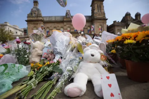 PA Media A teddy bear and flowers sit on the ground in a memorial