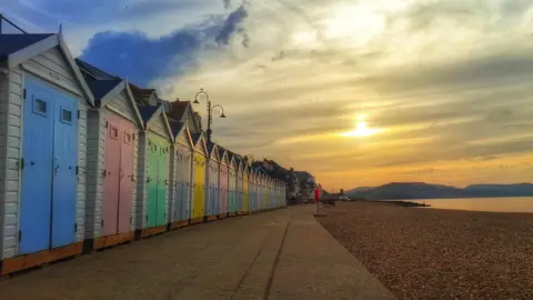 Ironsie A row of multi-coloured huts on a stony beach as the sun sets.