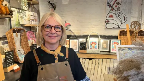 Lisa, a blonde lady with glasses stood in her art studio, surrounded by paintings and sculptures