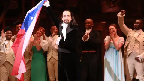 Getty Images Lin-Manuel Miranda standing on-stage at the end of a performance of Hamilton, holding the Puerto Rico flag