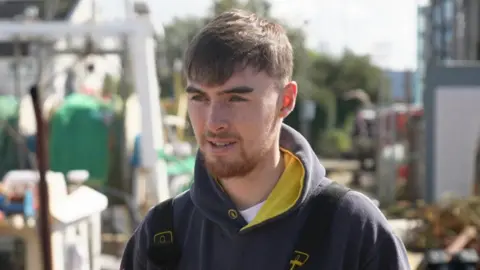 Joe Lynch looking to the left and wearing a blue hooded jacket. He has short brown hair and a short beard. 