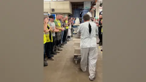 A man wearing white overalls is wheeling a trolley of pottery through a factory, as colleagues either side of him clap