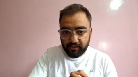 A man wearing a white t-shirt with spectacles looks into the camera. 